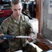 A U.S. Army soldier named Breault, wearing Operational Camouflage Pattern (OCP) uniform, is handling a small electronic device connected to multiple cables inside what appears to be a military operations vehicle or command post. He appears to be inspecting or connecting communications or electronic equipment. The setting suggests a field operations or training environment.