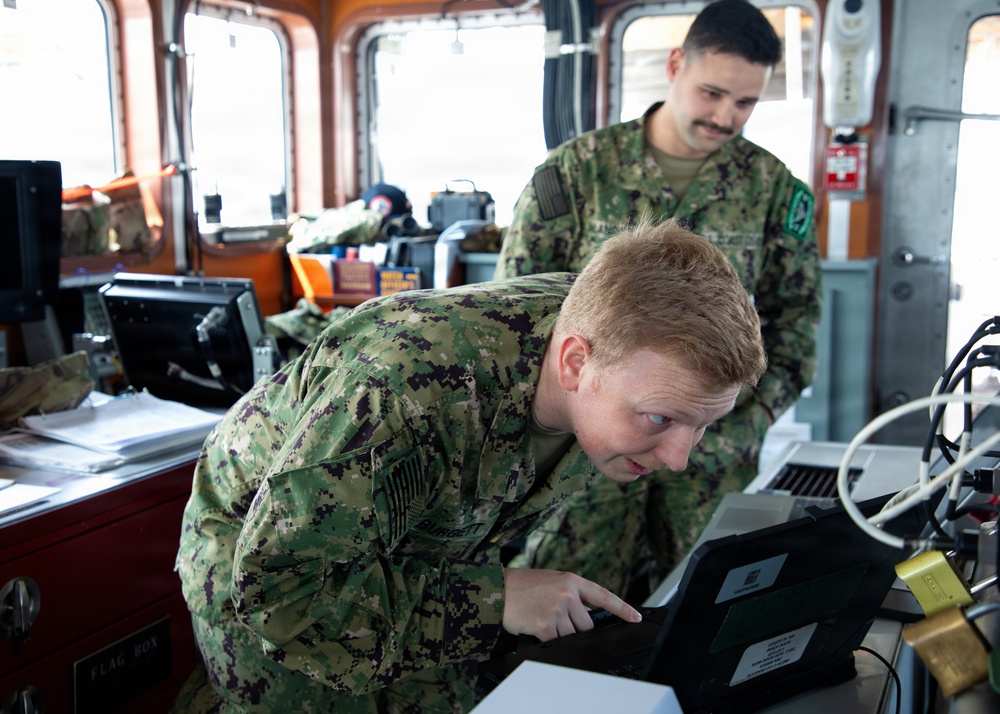 Two military personnel in camouflage uniforms are working inside what appears to be a vessel's bridge or command station. The foreground individual is leaning over and pointing at a ruggedized laptop/tablet device, while a second person stands behind observing. The setting contains multiple electronic displays, cables, and operational equipment consistent with a naval vessel's control room.