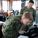 Two military personnel in camouflage uniforms are working inside what appears to be a vessel's bridge or command station. The foreground individual is leaning over and pointing at a ruggedized laptop/tablet device, while a second person stands behind observing. The setting contains multiple electronic displays, cables, and operational equipment consistent with a naval vessel's control room.