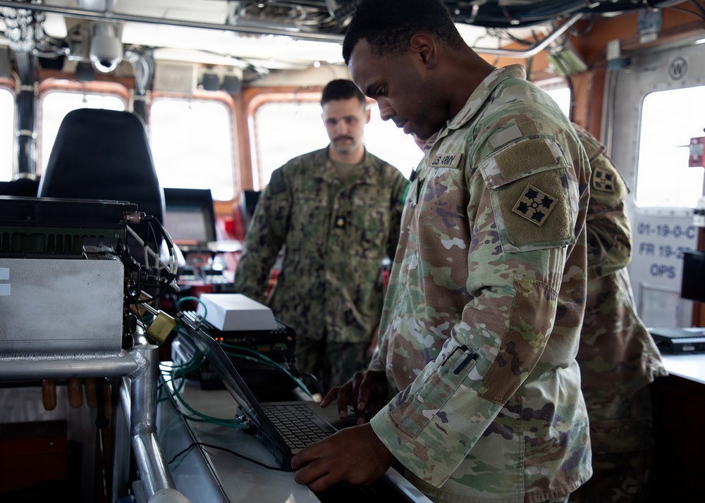 Two military personnel are working inside what appears to be a ship's bridge or command center. A U.S. Army soldier in the foreground is operating a laptop connected to various electronic equipment, while a second service member in the background observes. The setting contains multiple electronic systems, cables, and navigation-related equipment.