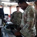 Two military personnel are working inside what appears to be a ship's bridge or command center. A U.S. Army soldier in the foreground is operating a laptop connected to various electronic equipment, while a second service member in the background observes. The setting contains multiple electronic systems, cables, and navigation-related equipment.