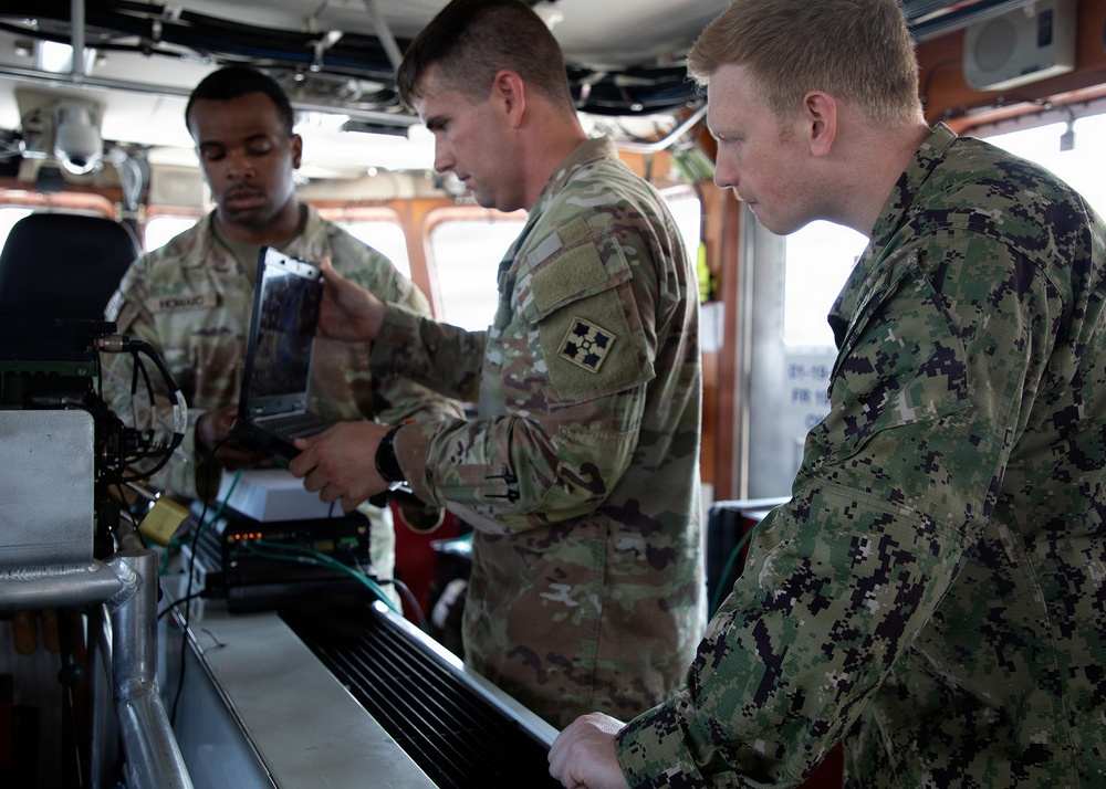 Three military personnel in different camouflage uniforms are gathered around electronic equipment aboard what appears to be a vessel's bridge or command area. Two soldiers in U.S. Army OCP (Operational Camouflage Pattern) uniforms and one sailor in U.S. Navy NWU (Naval Working Uniform) Type III are examining and operating what appears to be a laptop and associated hardware. The setting suggests a joint-service military operation or training exercise aboard a ship.