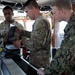 Three military personnel in different camouflage uniforms are gathered around electronic equipment aboard what appears to be a vessel's bridge or command area. Two soldiers in U.S. Army OCP (Operational Camouflage Pattern) uniforms and one sailor in U.S. Navy NWU (Naval Working Uniform) Type III are examining and operating what appears to be a laptop and associated hardware. The setting suggests a joint-service military operation or training exercise aboard a ship.