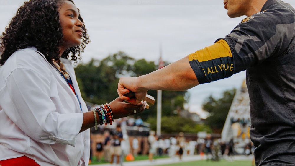 A woman in a white shirt and colorful bead bracelets is fist-bumping or exchanging a small object with a person wearing a dark athletic jersey with gold accents. The interaction appears to be taking place on or near a sports field, with a crowd of people visible in the blurred background. The mood appears positive and celebratory.