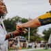 A woman in a white shirt and colorful bead bracelets is fist-bumping or exchanging a small object with a person wearing a dark athletic jersey with gold accents. The interaction appears to be taking place on or near a sports field, with a crowd of people visible in the blurred background. The mood appears positive and celebratory.