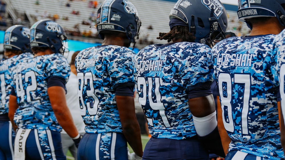 Old Dominion University (ODU) football players are shown from behind wearing special blue digital camouflage uniforms, likely for a military appreciation game. The jerseys display 'LTC SHAH' on the back instead of player names, suggesting a tribute to a military figure. Players wearing numbers 42, 83, 24, and 87 are visible standing on the sideline.