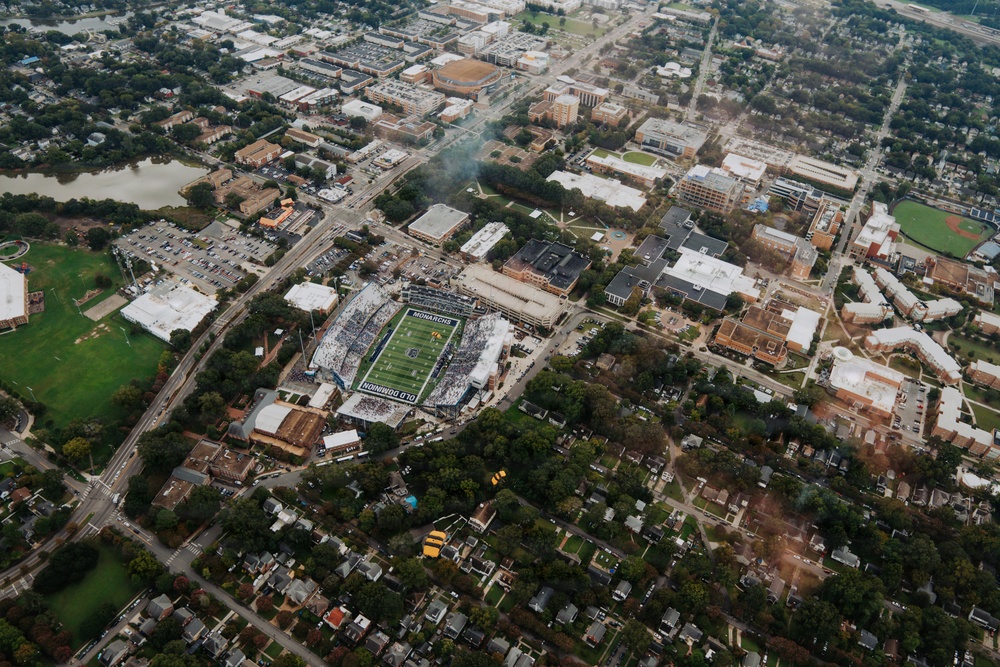 An aerial photograph of Old Dominion University's campus in Norfolk, Virginia, captured during a football game in progress at S.B. Ballard Stadium. The stadium, clearly labeled 'OLD DOMINION' and 'MONARCHS,' is filled with spectators and sits embedded within a dense urban environment. The surrounding area includes university buildings, residential neighborhoods, parking lots, and green spaces.