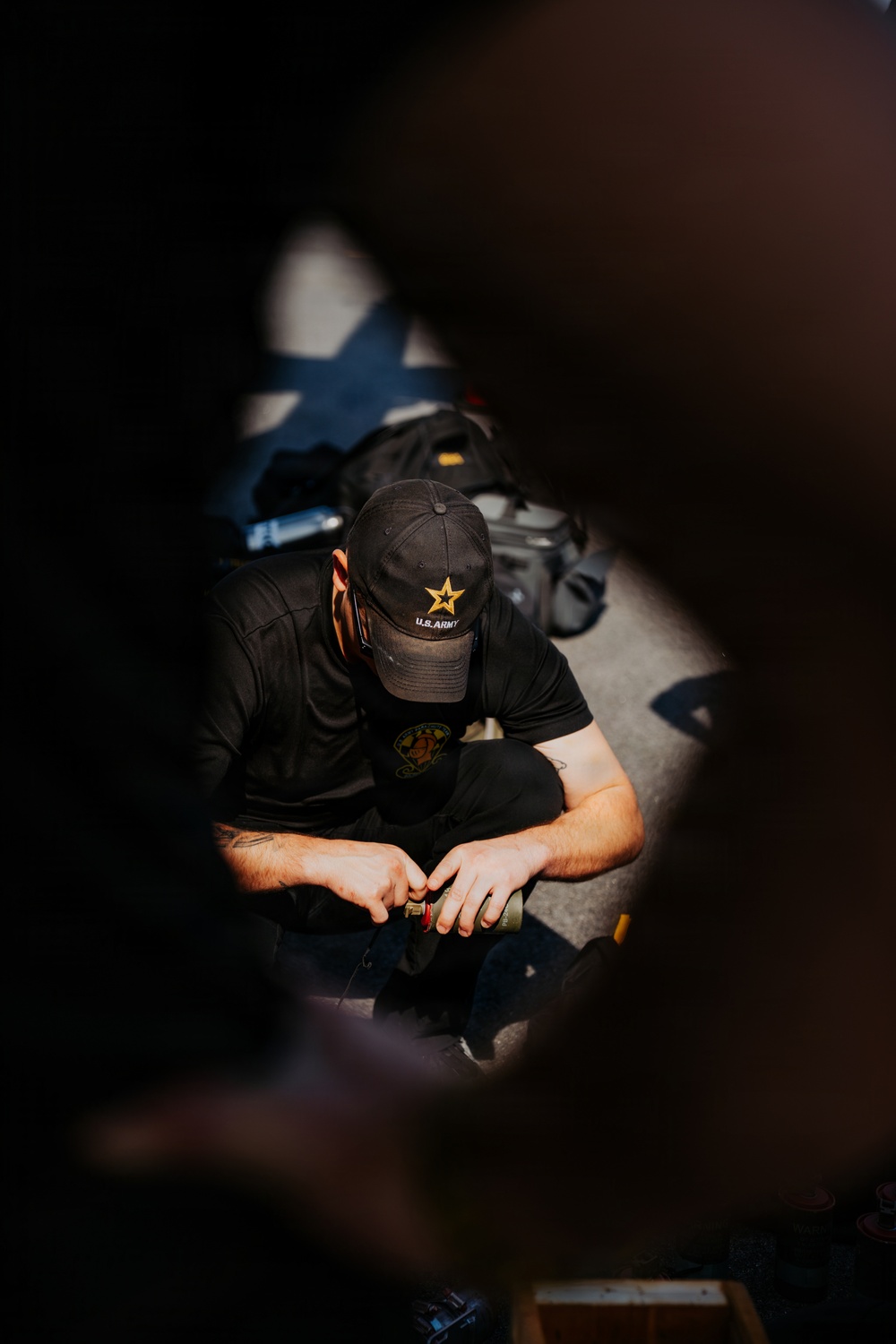 A person wearing a black U.S. Army branded baseball cap and black t-shirt is seated on the ground, viewed from above through a narrow opening or gap. They appear to be handling a small object with both hands. The image is shot with a shallow depth of field, with dark foreground elements framing the subject.