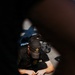 A person wearing a black U.S. Army branded baseball cap and black t-shirt is seated on the ground, viewed from above through a narrow opening or gap. They appear to be handling a small object with both hands. The image is shot with a shallow depth of field, with dark foreground elements framing the subject.