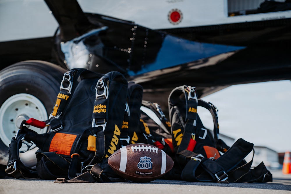 The image shows U.S. Army Golden Knights parachute rigs laid out on a tarmac surface next to an aircraft, alongside an ODU (Old Dominion University) Team Issue football. The equipment suggests a promotional or ceremonial event combining military skydiving with college football. The composition appears to be a staged publicity or pre-event photograph.