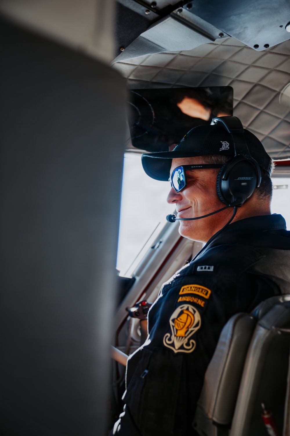 A male pilot is seated inside a small aircraft cockpit, wearing a Bose aviation headset, mirrored sunglasses, and a black cap. His dark jacket displays military-style patches including 'Ranger,' 'Airborne,' and what appears to be an 18th Airborne Corps or similar unit insignia. He is smiling slightly and looking forward through the cockpit window.