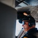 A male pilot is seated inside a small aircraft cockpit, wearing a Bose aviation headset, mirrored sunglasses, and a black cap. His dark jacket displays military-style patches including 'Ranger,' 'Airborne,' and what appears to be an 18th Airborne Corps or similar unit insignia. He is smiling slightly and looking forward through the cockpit window.