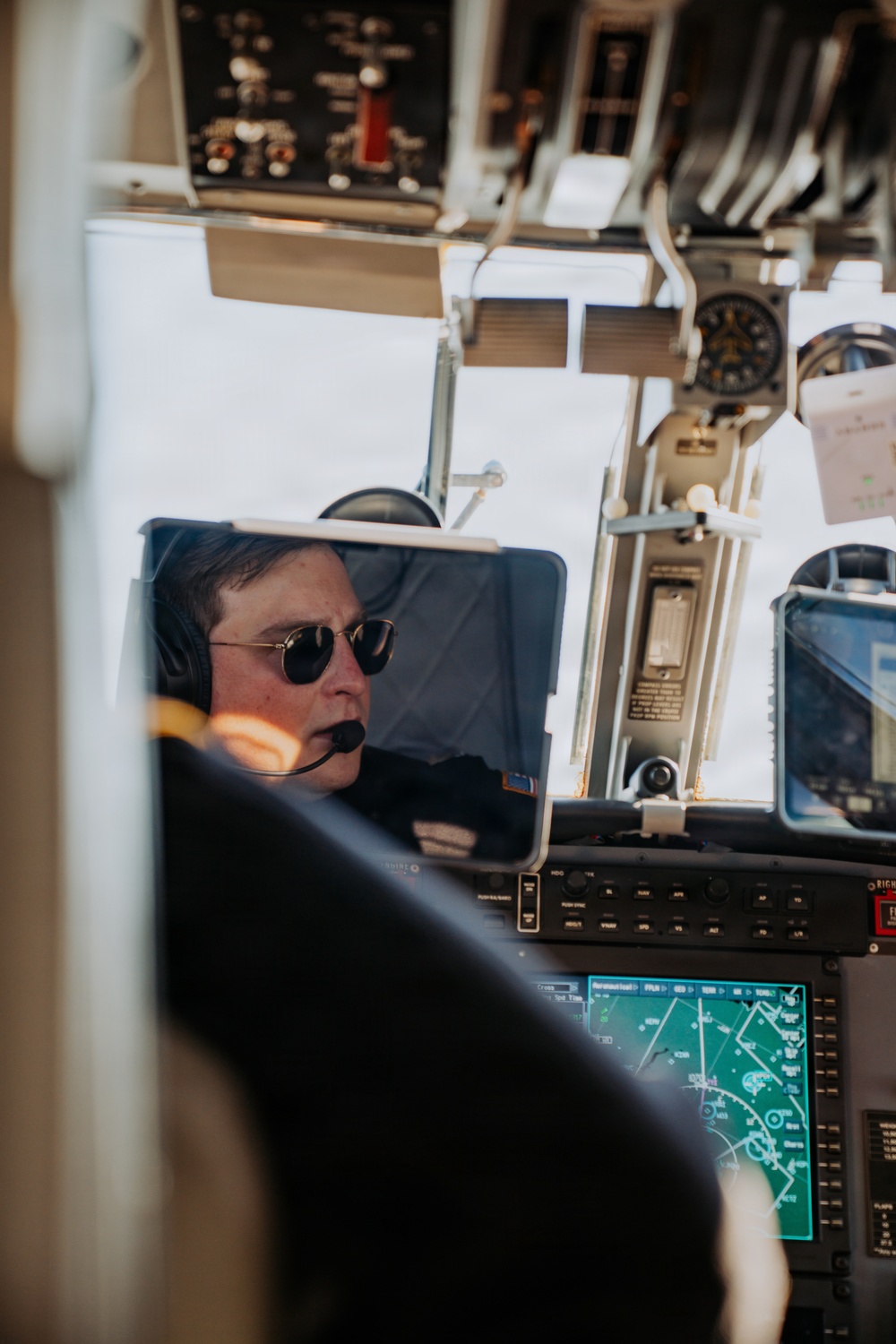 A pilot is seated in an aircraft cockpit, wearing aviator sunglasses and a aviation headset with a boom microphone. The cockpit features a mix of traditional analog instruments and modern digital avionics displays, including what appears to be a navigation/moving map display. The image is taken from behind and to the side, likely from a passenger or observer position.