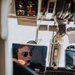 A pilot is seated in an aircraft cockpit, wearing aviator sunglasses and a aviation headset with a boom microphone. The cockpit features a mix of traditional analog instruments and modern digital avionics displays, including what appears to be a navigation/moving map display. The image is taken from behind and to the side, likely from a passenger or observer position.