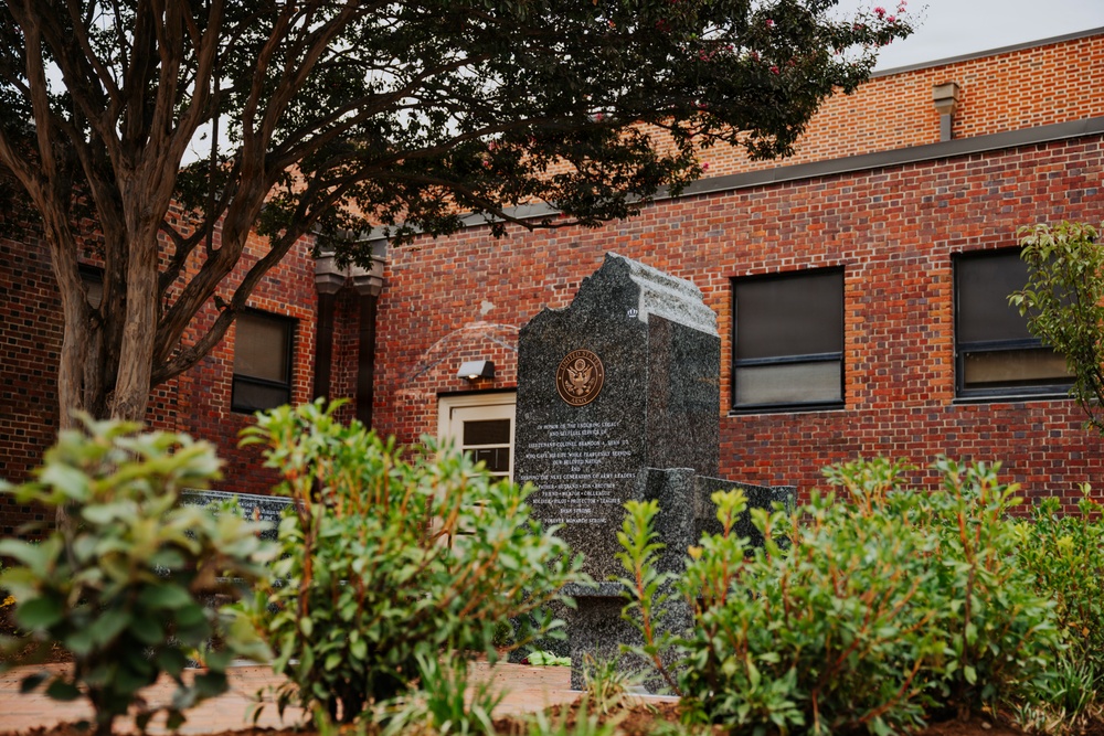 The image shows a dark granite memorial monument situated in a landscaped garden area in front of a red brick building. The monument features an embossed seal, likely the United States seal, along with inscribed text that is partially legible. The setting appears to be a public or governmental space, surrounded by shrubs and trees.