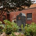 The image shows a dark granite memorial monument situated in a landscaped garden area in front of a red brick building. The monument features an embossed seal, likely the United States seal, along with inscribed text that is partially legible. The setting appears to be a public or governmental space, surrounded by shrubs and trees.