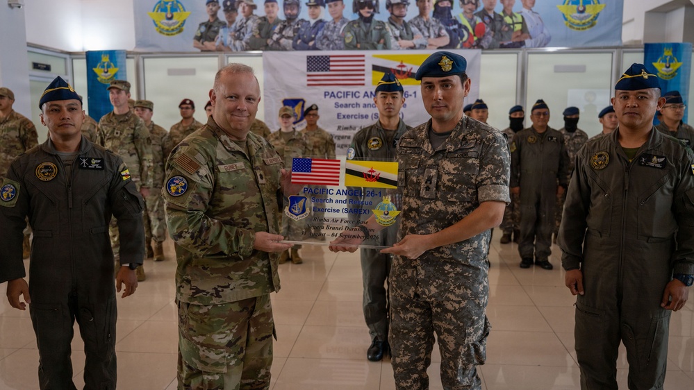 Two military officers, one American and one Bruneian, are holding a commemorative plaque for 'Pacific Angel 26-1 Search and Rescue Exercise (SAREX)' held at Rimba Air Force Base, Negara Brunei Darussalam. The ceremony appears to be a formal exchange or closing ceremony for a joint military exercise between the United States and Brunei. Personnel from both nations are assembled in formation behind the two principal officers.