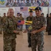 Two military officers, one American and one Bruneian, are holding a commemorative plaque for 'Pacific Angel 26-1 Search and Rescue Exercise (SAREX)' held at Rimba Air Force Base, Negara Brunei Darussalam. The ceremony appears to be a formal exchange or closing ceremony for a joint military exercise between the United States and Brunei. Personnel from both nations are assembled in formation behind the two principal officers.