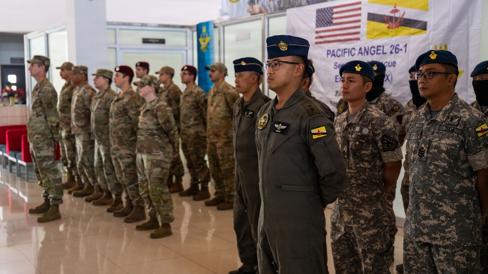 Military personnel from the United States and Brunei are standing in formation at a joint exercise called 'Pacific Angel 26-1,' a Search and Rescue Exercise (SAREX). The banner in the background displays both the American and Bruneian flags alongside the exercise name. Personnel are wearing a mix of US military camouflage uniforms and Bruneian military uniforms including flight suits and digital camouflage.