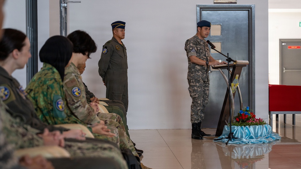 A military ceremony or briefing is taking place indoors, with a uniformed officer speaking at a podium equipped with a microphone. Several military personnel in various camouflage uniforms are seated as an audience, while another officer stands at attention nearby. The setting appears formal, with decorative flowers arranged at the base of the podium.