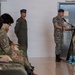 A military ceremony or briefing is taking place indoors, with a uniformed officer speaking at a podium equipped with a microphone. Several military personnel in various camouflage uniforms are seated as an audience, while another officer stands at attention nearby. The setting appears formal, with decorative flowers arranged at the base of the podium.