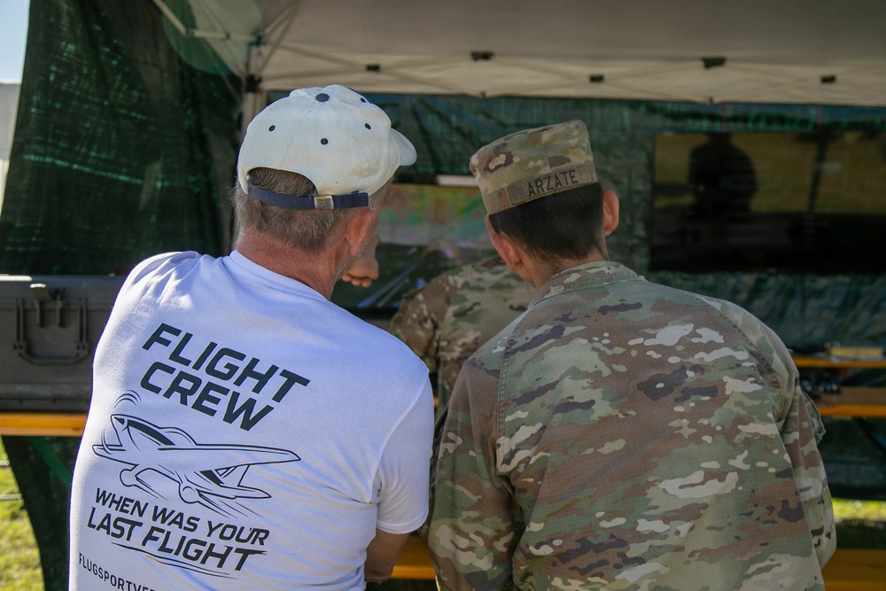 Two individuals are seen from behind, standing under a canopy tent and appearing to review something together. One person wears a white 'Flight Crew' t-shirt and white baseball cap, while the other is dressed in military camouflage (OCP/MultiCam) uniform with a name tape reading 'ARZATE'. The setting appears to be an outdoor event or field operation.