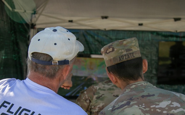 Two individuals are seen from behind, standing under a canopy tent and appearing to review something together. One person wears a white 'Flight Crew' t-shirt and white baseball cap, while the other is dressed in military camouflage (OCP/MultiCam) uniform with a name tape reading 'ARZATE'. The setting appears to be an outdoor event or field operation.