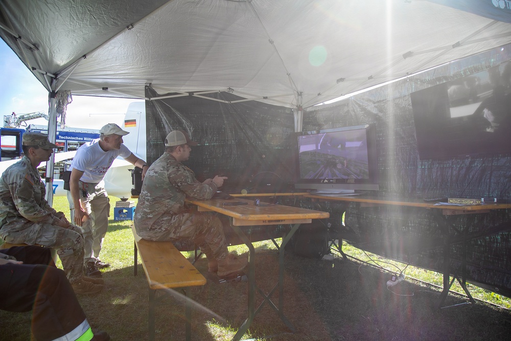 A group of individuals, including at least two in military camouflage uniforms, are gathered under a white canopy tent in an outdoor setting. They appear to be operating or observing equipment displayed on a monitor, possibly related to drone or UAV operations. A German flag is visible on a backdrop in the background, suggesting an international military or civil defense event.