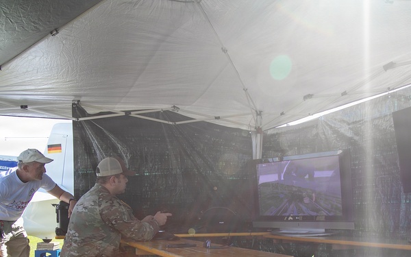 A group of individuals, including at least two in military camouflage uniforms, are gathered under a white canopy tent in an outdoor setting. They appear to be operating or observing equipment displayed on a monitor, possibly related to drone or UAV operations. A German flag is visible on a backdrop in the background, suggesting an international military or civil defense event.