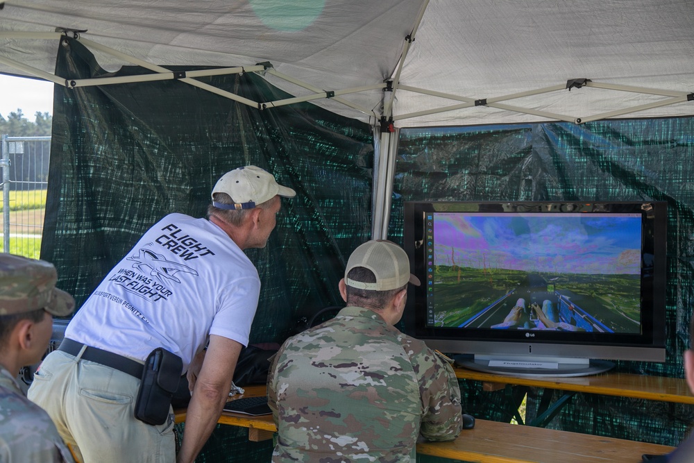 Several individuals are gathered under a portable canopy tent observing a large LG monitor displaying what appears to be a drone or aerial vehicle feed. One person in a white 'Flight Crew' t-shirt is leaning over to observe, while a person in military camouflage uniform sits at the table operating equipment. The setup suggests a field drone operations or UAV demonstration station.