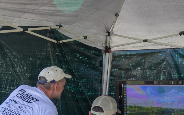 Several individuals are gathered under a portable canopy tent observing a large LG monitor displaying what appears to be a drone or aerial vehicle feed. One person in a white 'Flight Crew' t-shirt is leaning over to observe, while a person in military camouflage uniform sits at the table operating equipment. The setup suggests a field drone operations or UAV demonstration station.