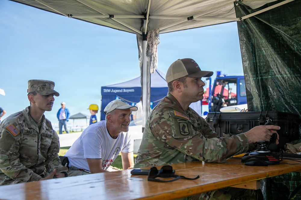 Two U.S. Army soldiers in OCP (Operational Camouflage Pattern) uniforms and a civilian are gathered under a white canopy tent at what appears to be an outdoor public event or demonstration. One soldier is operating what appears to be a drone control station or similar military equipment mounted in a hard-shell case. A banner in the background reads 'THW-Jugend Bayern', suggesting this event is taking place in Bavaria, Germany.