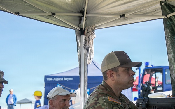 Two U.S. Army soldiers in OCP (Operational Camouflage Pattern) uniforms and a civilian are gathered under a white canopy tent at what appears to be an outdoor public event or demonstration. One soldier is operating what appears to be a drone control station or similar military equipment mounted in a hard-shell case. A banner in the background reads 'THW-Jugend Bayern', suggesting this event is taking place in Bavaria, Germany.