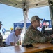 Two U.S. Army soldiers in OCP (Operational Camouflage Pattern) uniforms and a civilian are gathered under a white canopy tent at what appears to be an outdoor public event or demonstration. One soldier is operating what appears to be a drone control station or similar military equipment mounted in a hard-shell case. A banner in the background reads 'THW-Jugend Bayern', suggesting this event is taking place in Bavaria, Germany.