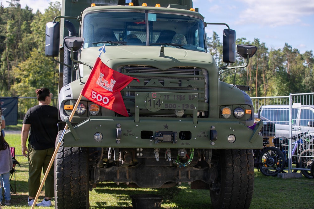 A front-facing view of a military olive-drab Mack truck displayed at what appears to be an outdoor public event or show. The vehicle bears U.S. Army markings and a red flag with '500' attached to the front. Spectators and other vehicles are visible in the background.