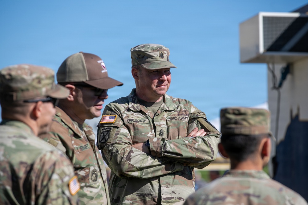 A group of U.S. Army soldiers in OCP (Operational Camouflage Pattern) uniforms are gathered outdoors in what appears to be a military setting. The central figure, identified by his name tape as 'CHRZANOWSKI,' is a senior NCO wearing a Sergeant Major rank insignia, standing with arms crossed and smiling. The soldiers appear to be engaged in a casual conversation or briefing.