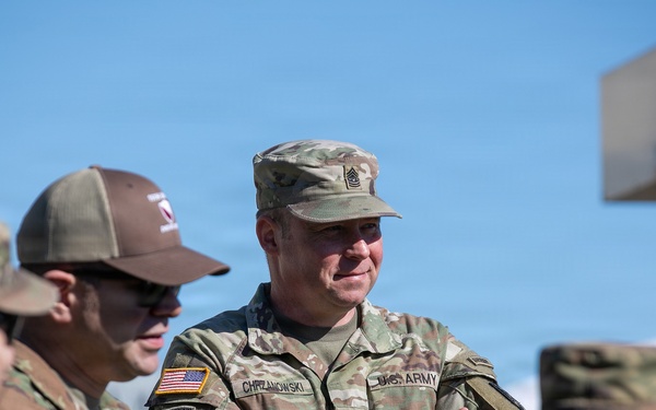A group of U.S. Army soldiers in OCP (Operational Camouflage Pattern) uniforms are gathered outdoors in what appears to be a military setting. The central figure, identified by his name tape as 'CHRZANOWSKI,' is a senior NCO wearing a Sergeant Major rank insignia, standing with arms crossed and smiling. The soldiers appear to be engaged in a casual conversation or briefing.