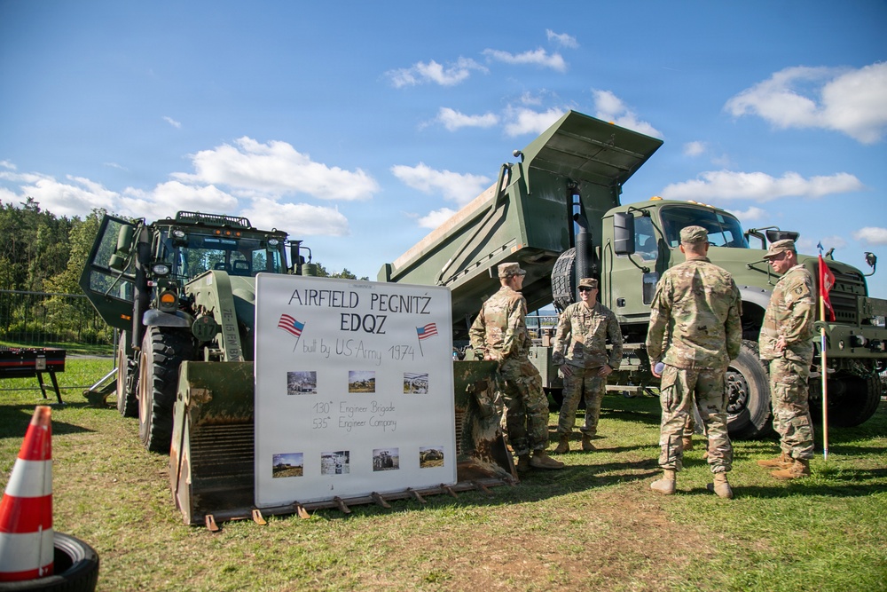 A group of U.S. Army soldiers in OCP camouflage uniforms are gathered around military engineering equipment at an outdoor display. A whiteboard sign identifies the display as related to 'Airfield Pegnitz EDQZ,' built by the U.S. Army in 1974, associated with the 130th Engineer Brigade and 535th Engineer Company. The scene appears to be a static equipment demonstration or open house event on a grassy field.