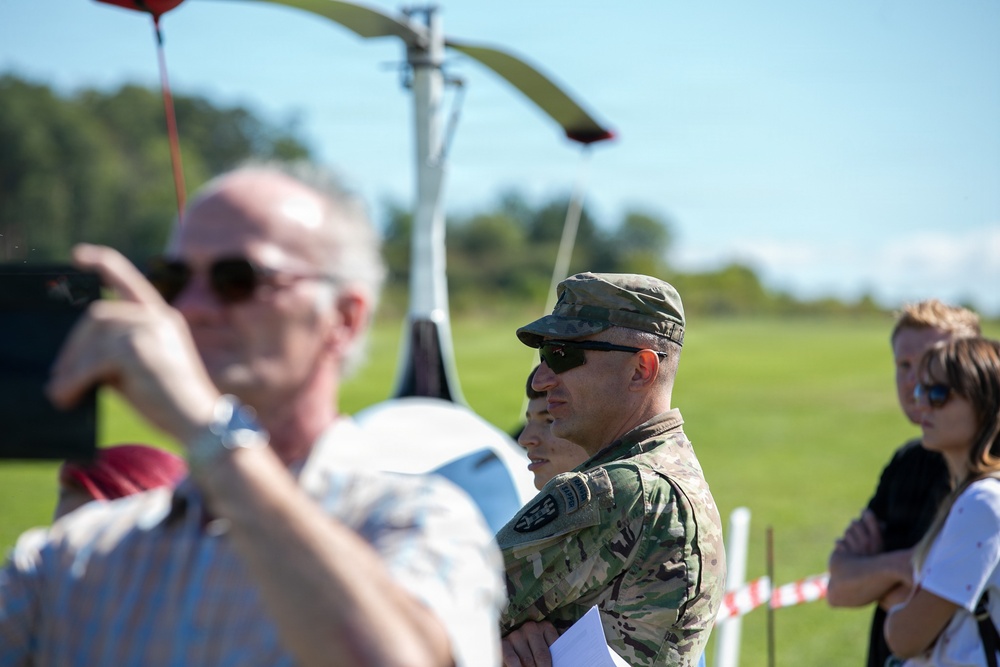 A group of people are gathered outdoors near what appears to be a small helicopter or autogyro on a grassy airfield. A military personnel in camouflage uniform and patrol cap is prominently visible in the center, alongside civilians. The scene suggests an airshow, demonstration, or military aviation event.