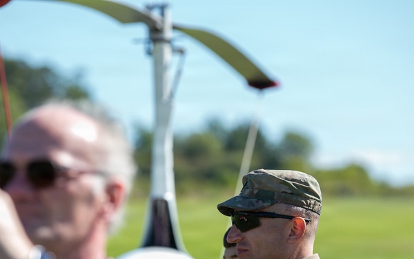 A group of people are gathered outdoors near what appears to be a small helicopter or autogyro on a grassy airfield. A military personnel in camouflage uniform and patrol cap is prominently visible in the center, alongside civilians. The scene suggests an airshow, demonstration, or military aviation event.