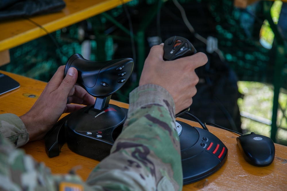 A person wearing military camouflage (multicam/OCP pattern) uniform is operating two joystick controllers on a wooden table in an outdoor setting. The setup appears to be a drone or UAV ground control station, with dual joysticks and a computer mouse visible. The environment suggests a field operation or training exercise.