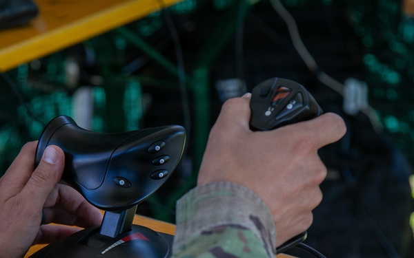 A person wearing military camouflage (multicam/OCP pattern) uniform is operating two joystick controllers on a wooden table in an outdoor setting. The setup appears to be a drone or UAV ground control station, with dual joysticks and a computer mouse visible. The environment suggests a field operation or training exercise.