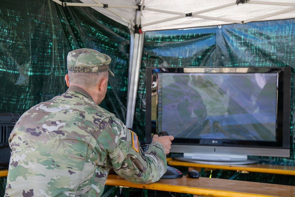 A U.S. Army soldier identified by name tape as 'TABACCHI' is seated at a field workstation under a canopy tent, operating what appears to be a drone or UAV ground control system. The soldier is using a handheld controller while viewing an aerial or satellite-feed display on an LG monitor. The setup suggests a field exercise or operational drone control station.