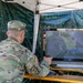 A U.S. Army soldier identified by name tape as 'TABACCHI' is seated at a field workstation under a canopy tent, operating what appears to be a drone or UAV ground control system. The soldier is using a handheld controller while viewing an aerial or satellite-feed display on an LG monitor. The setup suggests a field exercise or operational drone control station.