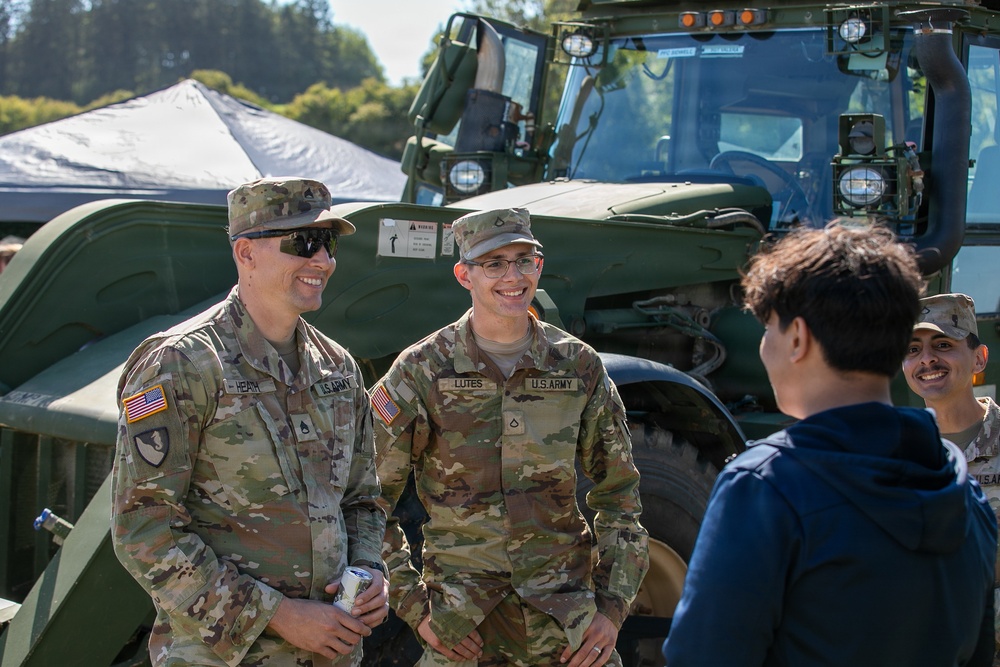 Two U.S. Army soldiers in OCP (Operational Camouflage Pattern) uniforms are engaged in a friendly conversation with a civilian, likely at a public military outreach or recruitment event. Name tapes visible on the uniforms read 'HEATH' and 'LUTES', both bearing U.S. Army insignia. A large olive-drab military vehicle is visible in the background along with a third soldier partially visible on the right.
