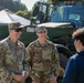 Two U.S. Army soldiers in OCP (Operational Camouflage Pattern) uniforms are engaged in a friendly conversation with a civilian, likely at a public military outreach or recruitment event. Name tapes visible on the uniforms read 'HEATH' and 'LUTES', both bearing U.S. Army insignia. A large olive-drab military vehicle is visible in the background along with a third soldier partially visible on the right.