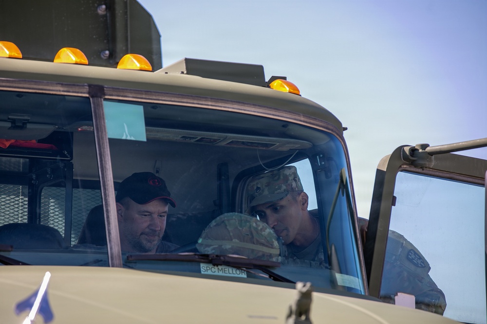 Two individuals are seated inside the cab of a military vehicle, with one wearing civilian clothing and a black cap, and the other in military camouflage uniform. A name placard reading 'SPC MELLOR' is visible on the vehicle's dashboard area. They appear to be engaged in conversation or reviewing something together.