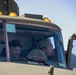 Two individuals are seated inside the cab of a military vehicle, with one wearing civilian clothing and a black cap, and the other in military camouflage uniform. A name placard reading 'SPC MELLOR' is visible on the vehicle's dashboard area. They appear to be engaged in conversation or reviewing something together.