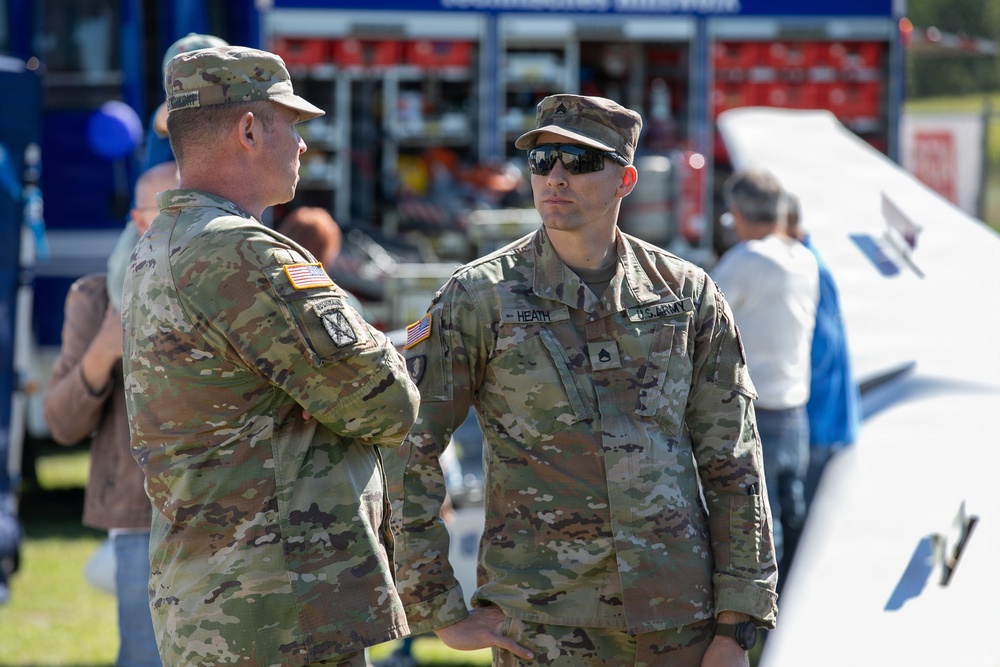 Two U.S. Army soldiers in Operational Camouflage Pattern (OCP) uniforms are engaged in conversation at what appears to be a public military event or community outreach. The soldier on the right is identified by his name tape as 'HEATH' and wears 'U.S. ARMY' chest tape along with sunglasses. The background shows emergency vehicles, civilians, and event infrastructure suggesting an outdoor public display.