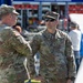 Two U.S. Army soldiers in Operational Camouflage Pattern (OCP) uniforms are engaged in conversation at what appears to be a public military event or community outreach. The soldier on the right is identified by his name tape as 'HEATH' and wears 'U.S. ARMY' chest tape along with sunglasses. The background shows emergency vehicles, civilians, and event infrastructure suggesting an outdoor public display.